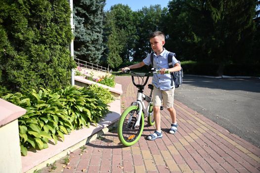 Full-length portrait of adorable child parking his bike at school entrance. Schoolboy coming back to school