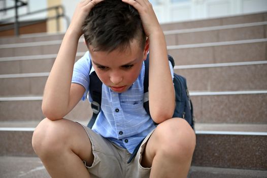 Frustrated worried schoolboy holding his head with his hands while sitting on the stairs near the school. Concept of learning difficulties, bullying, loneliness. Back to school