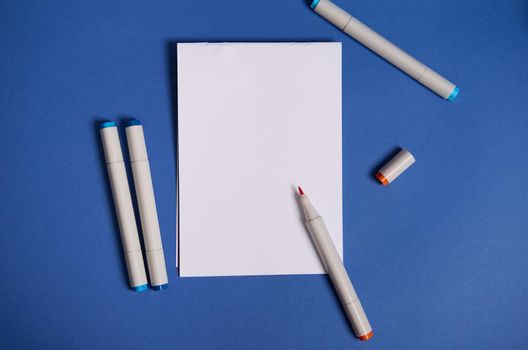 Dichromatic flat lay composition with watercolor markers or felt-tip pens with a white blank empty paper sheet with copy space, isolated on blue background.