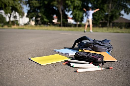 Scattered school supplies, pencil case, markers, workbooks falling out of an open backpack, lie on the asphalt of the schoolyard against the background of a blurred happy schoolboy raising his hands up