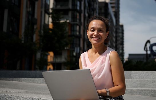 Close-up portrait of a cute business woman of mixed race ethnicity working on laptop sitting on the steps in the city and smiling looking at camera. Business, distant remote work and start-up concept