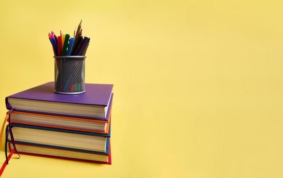 A metal pencil holder with school supplies on stacks of multicolored books. Teacher's Day concept, literary, knowledge, Back to School, education. Isolated on yellow background copy space