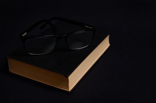 Monochrome composition of eyeglasses on a book in hard black cover, isolated over black background with space for text. Teacher's Day concept, Knowledge, literature ,reading, erudition