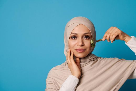 Beautiful Muslim woman in hijab massaging her face with jade roller. Isolated beauty portrait on blue background with space for text