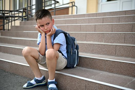 Bullying, loneliness, studying difficulties. Upset worried boy with backpack sitting on the stairs by school.