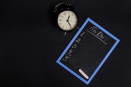 Top view of a chalk board with lettering list to do, with empty copy space and a vintage alarm clock. Isolated on black background. Planning concepts, time management, timetable and timing.