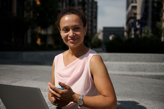 Confident close-up portrait of a beautiful mixed race business woman with laptop and smartphone looking at the camera sitting on steps on the background of urban tall buildings