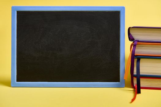 A blank chalkboard and multicolored books on yellow surface background with copy space for text. Teacher's Day concept, Knowledge, literature ,reading, erudition