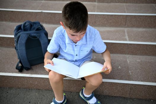 Elementary student doing homework sitting next to his school bag on the stairs of school establishment. Concept of back to school