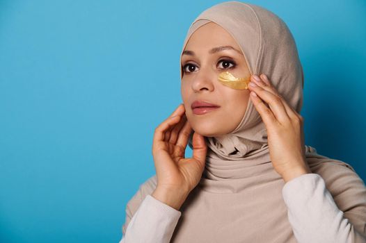 Closeup portrait of a Muslim woman in hijab using hydrogel eye patches, on blue background with copy space