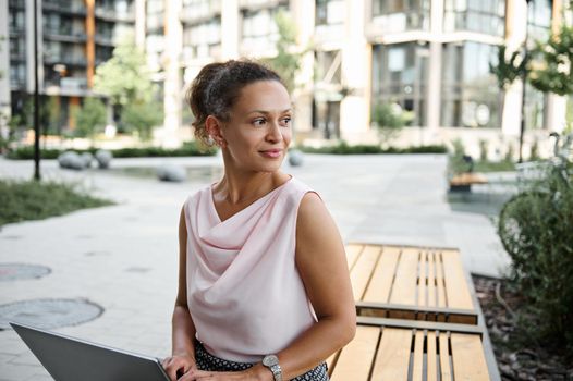 Attractive young mixed race business lady, woman freelancer working on laptop, looking away while sitting on wooden bench in city