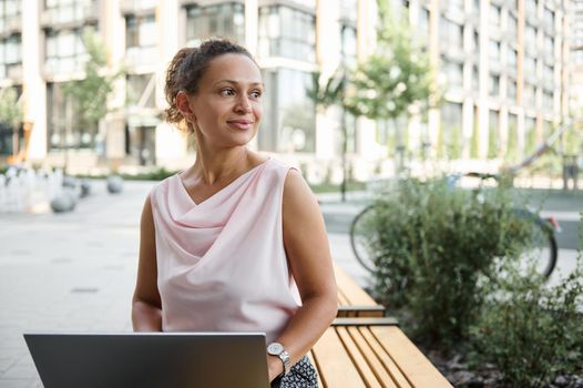 Attractive young African American woman freelancer working on laptop, looking away while sitting on wooden bench in city