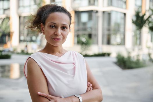 Close-up confident portrait of a successful middle aged business woman in casual attire posing to camera with crossed rams on the background of high buildings. Urban city backdrop