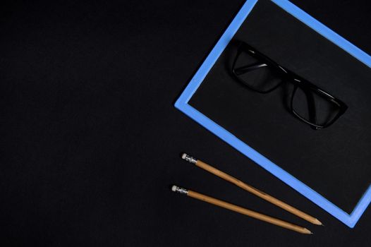 Flat lay of eyeglasses on an empty blank chalkboars and two wooden pencils on black surface background with coy space