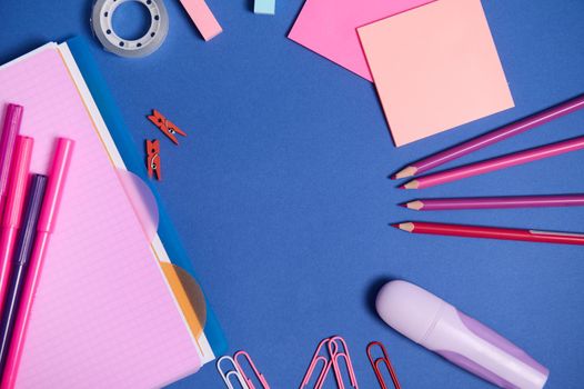 Assortment of school office supplies in pink shades, isolated on blue background, copy space. Copybook, felt tip pens, markers, paper clips,pencils and stationery accessories scattered in a circle