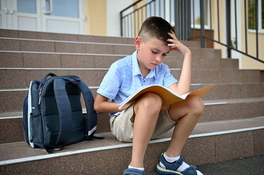 Puzzled elementary school student, pupil, school boy, doing his homework on a notebook while sitting on the stairs of a school institution.