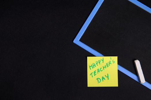 Flat lay of a pasted yellow note paper with inscription Happy Teacher's Day on an empty blank board with a chalk. Isolated on black background, copy space