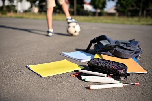 Scattered school supplies and workbooks falling out of an open backpack, lie on the asphalt of the schoolyard against the background of a boy's feet on a soccer ball. Recreation, back to school.
