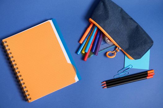Flat lay with copybook, organizer, colorful pencils and pencil case with falling out school supplies , in blue and orange contrast shadows isolated on blue background with copy space. High angle view