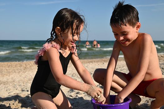 Happy brother and sister, friends,boy and girl filling the purple plastic toy bucket with wet sand and building sandy figures. Happy children playing on the beach at sea background, enjoying summer vacations. Summer holidays concept