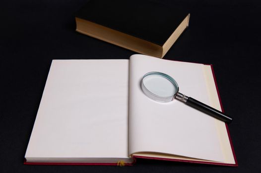 A magnifying glass, loupe, magnifier on an opened book in hard red cover, isolated over black background with copy space for text. Teacher's Day concept, Knowledge, literature ,reading, erudition