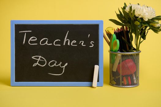 A stand for pens and pencils on the desktop on a yellow background with copy space. Holder with colored school supplies and aster flower and chalk board with lettering Teacher's Day