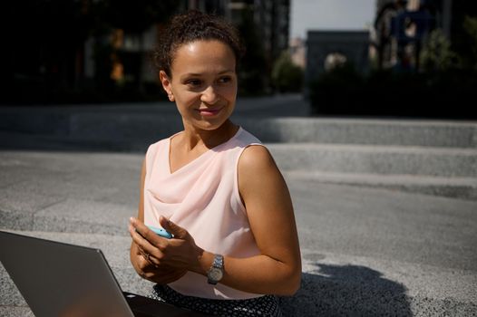 Confident portrait of a beautiful mixed race business woman with laptop and smartphone looking to the side sitting on steps on the background of urban tall buildings