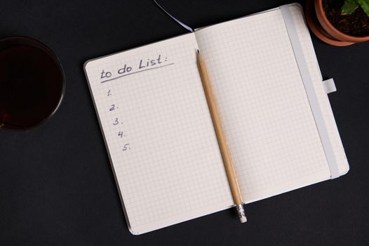 Wooden pencil on a notepad, organizer with list to do on blank white sheets, cup of coffee and potted succulent, on black background with copy space. Business, Organization, Time Management