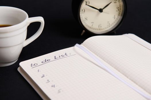 A cropped image of an opened organizer, notepad with list to do on blank white sheets, cup of coffee, alarm clock on black background with copy space. Business, Organization, Time Management concepts.