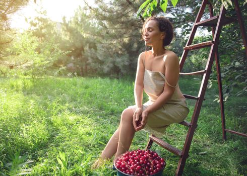 Beautiful mixed race woman in a linen dress sitting on a ladder next to a bucket of cherries in the orchard at sunset. Beautiful sun rays fall into the garden on a summer day
