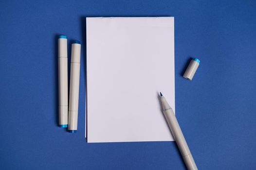 Two-color flat lay composition with watercolor markers or felt-tip pens with white blank sheet of paper with copy space isolated on blue background.