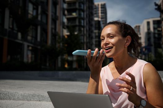 Adorable mixed race beautiful woman recording a voice message on smartphone while working on laptop sitting on steps on the buildings background. Business concept