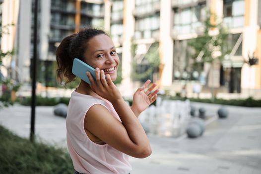 Attractive Hispanic young woman talking on mobile phone, cutely smiling with toothy smile while looking at the camera, standing on the urban buildings background. Business and communication concept