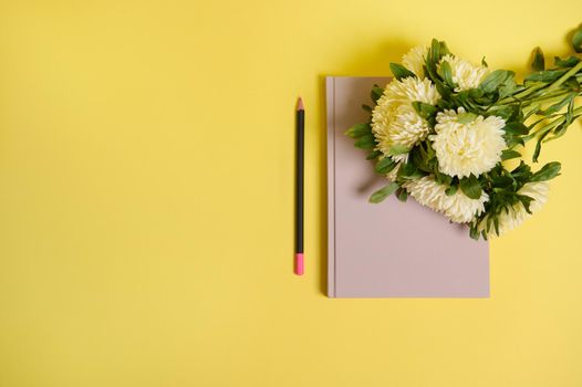 Flat lay of beautiful delicate bouquet of Autumn flowers, white asters lying down on the corner of a notepad organizer, next to a wooden color pencil, isolated over yellow background with copy space