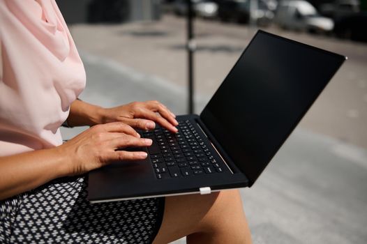 Close-up of female hands typing text on on laptop with blank black monitor screen with copy space for promotion on the urban background. Business concept