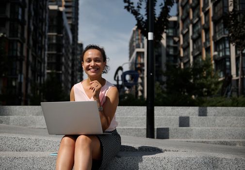 Front view of a cheerful pretty woman of mixed race ethnicity working on laptop sitting on the steps in the city and smiling looking at camera. Business, distant remote work and start-up concept