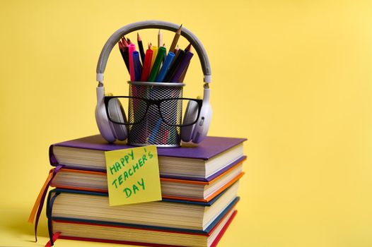 Composition from a stack of colorful books pencils glasses wireless headphones on a yellow background with copy space. Back to school concepts. Teachers Day.