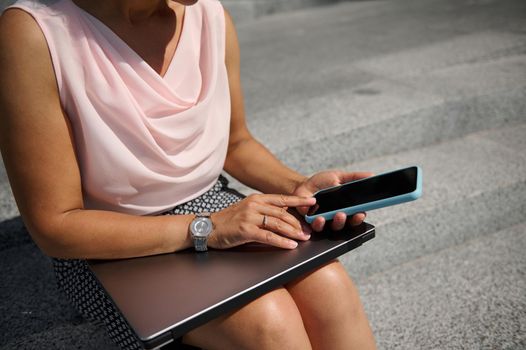 Close-up. Focus on a woman's hand with stylish hand watch, holding a smartphone , swiping mobile application, sitting on steps with a laptop. Business, freelance, online and distant work concept