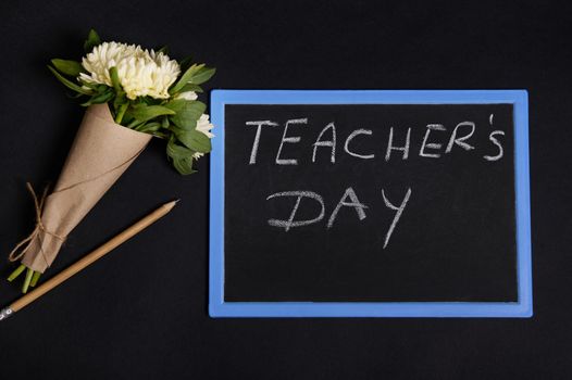 A wooden pencil and beautiful bouquet of asters flowers in craft wrapping paper lying down next to a chalk board with lettering Teacher's Day. Flat lay on black background with copy space