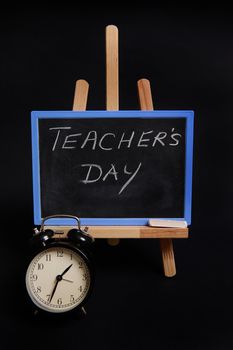 Close-up of a chalk blackboard with lettering Teacher's Day, standing on a wooden table easel, next to black alarm clock, isolated on black background with copy space.