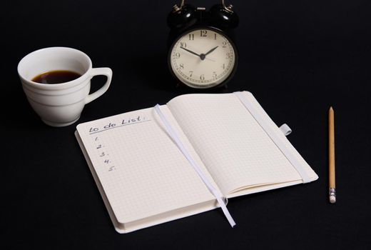 Opened organizer, notepad with list to do on blank white sheets, wooden pencil, cup of coffee and alarm clock on black background with copy space. Business, Organization, Time Management concepts.