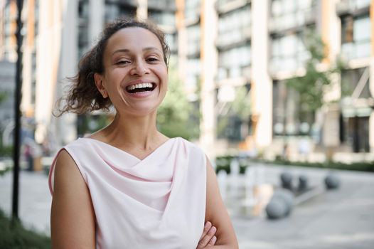 Attractive mixed race Hispanic, African American woman smiling with toothy smile standing on the urban buildings background. Confident portrait of a beautiful young business woman