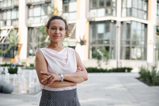 Confident business portrait of a successful middle aged business woman in casual attire posing to camera with crossed rams on the background of high buildings. Urban city background