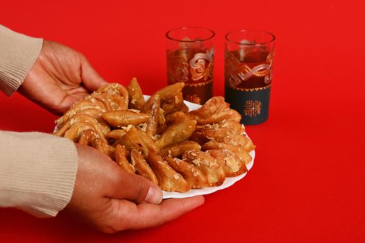 Close-up of hands serving a plate of delicious oriental traditional Moroccan sweet dessert and mint tea in a beautiful Arabic style glass. Isolated on red background with copy space