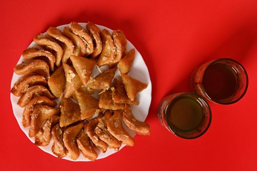 Flat composition with oriental moroccan dessert on plate next to two glasses in arabic style on red background with space for text. Arabic traditional oriental sweets on the festive table