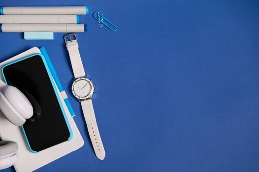 Flat lay of headphones, smartphone in blue case on white diary , a hand watch, markers and paper clips isolated on blue background with copy space. Top view of white and blue office school supplies
