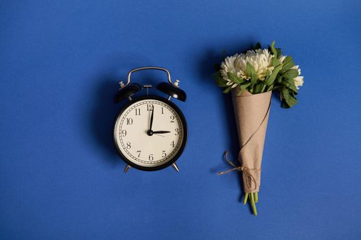 Flat lay composition of a beautiful bouquet of asters in craft paper and an alarm clock, lying on a blue background with space for text. Teachers' Day and Back to School Concepts