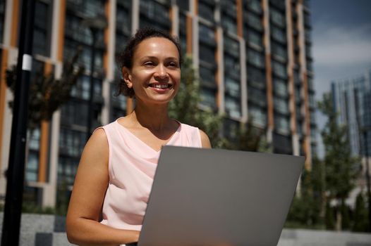 Happy middle aged business woman of mixed race ethnicity , office worker, employee smiling looking away, using laptop, sitting on steps on the high urban, city buildings background