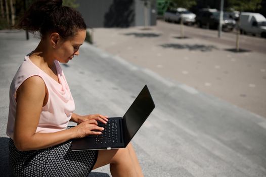 Side portrait of busy freelance African American woman, start-uper , office worker using laptop sitting on steps during coffee break. Office worker on lunch break in urban background