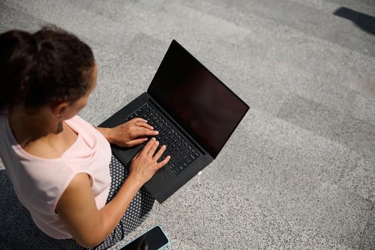 High angle view of a woman sitting on steps and typing text on on laptop with blank black monitor screen with copy space for advertising on the urban background. Business concept